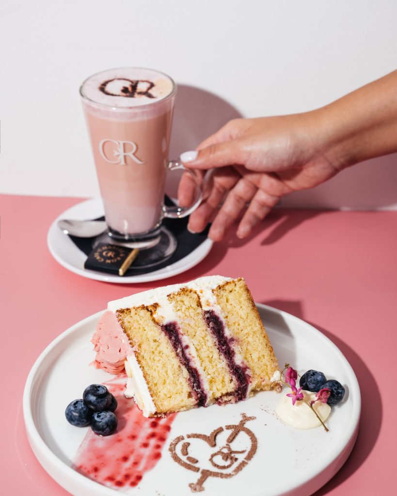 Layered cake with berry filling and cream, scoop of pink ice cream, blueberries, decorated cappuccino with "CR" art, hand on glass mug.